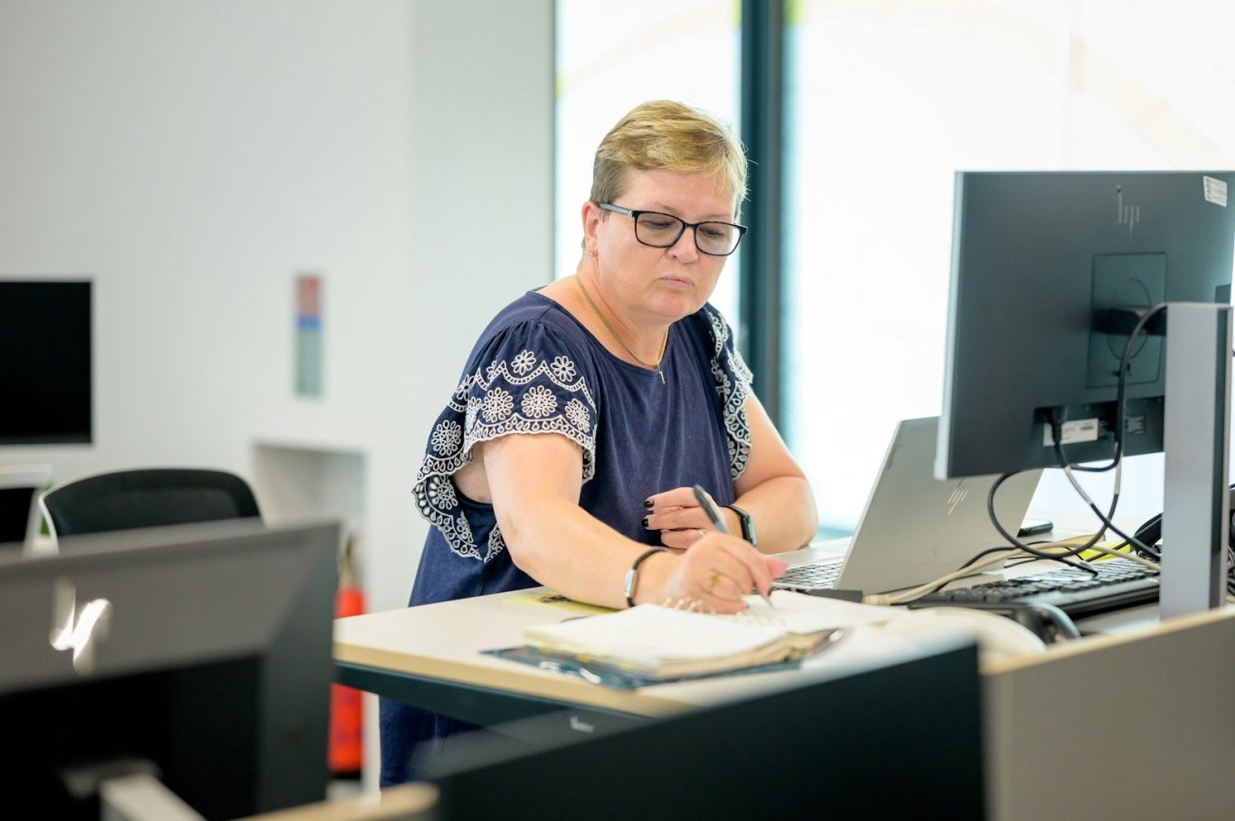 Image shows a woman sitting at a desk, writing in a notebook in front of her computer. Her expression is focused